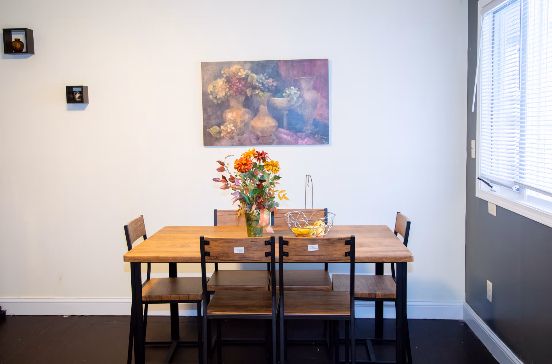 Dining area with a wooden table, six chairs, a floral centerpiece, and a painting on the wall.