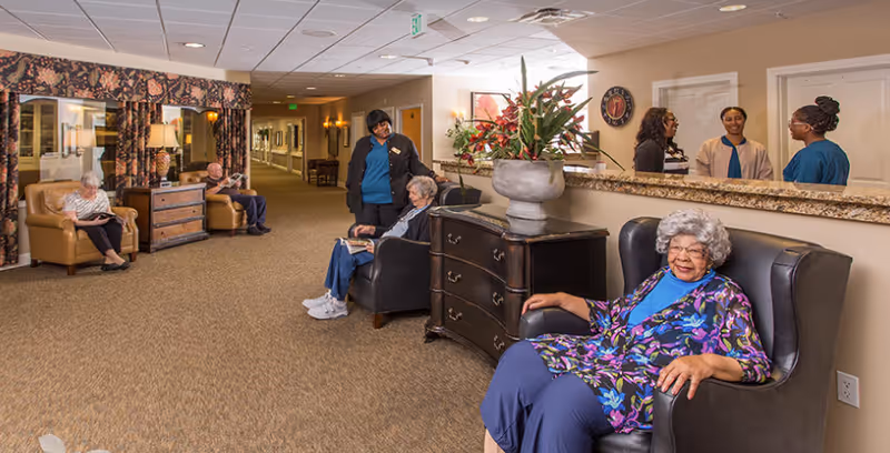 A spacious assisted living facility common area with several elderly residents seated comfortably in armchairs. A staff member is interacting with one resident, while three other staff members are conversing near a reception desk with a large floral arrangement. The area is warmly decorated with carpeted floors, lamps, and framed artwork.