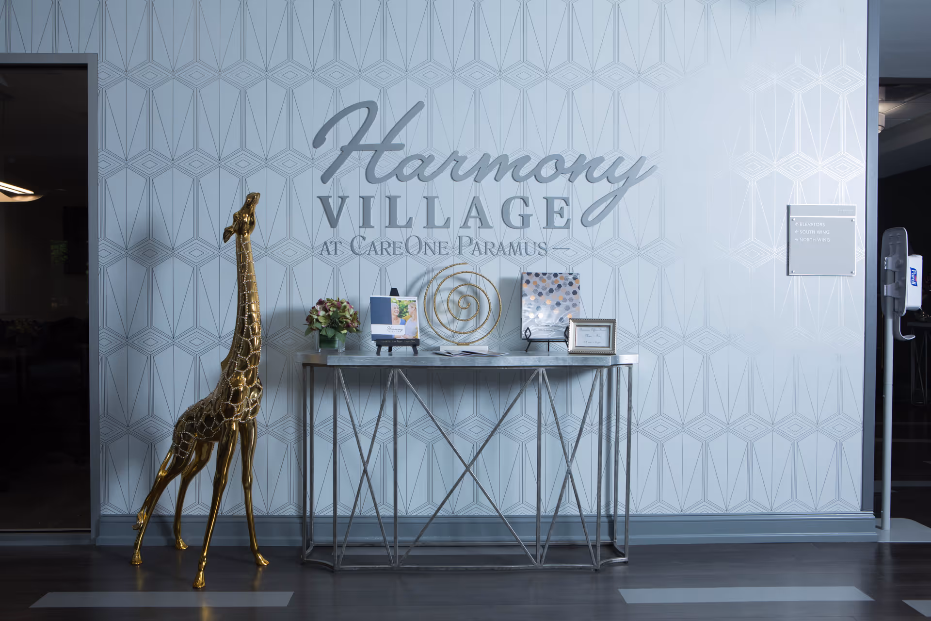 A modern interior wall with the text 'Harmony Village at CareOne Paramus' displayed prominently. In front of the wall is a metallic console table with decorative items including a small flower arrangement, framed pictures, and a spiral sculpture. To the left of the table stands a tall, golden giraffe statue. The floor is dark wood, and there is a hand sanitizer dispenser mounted on the right side of the wall.