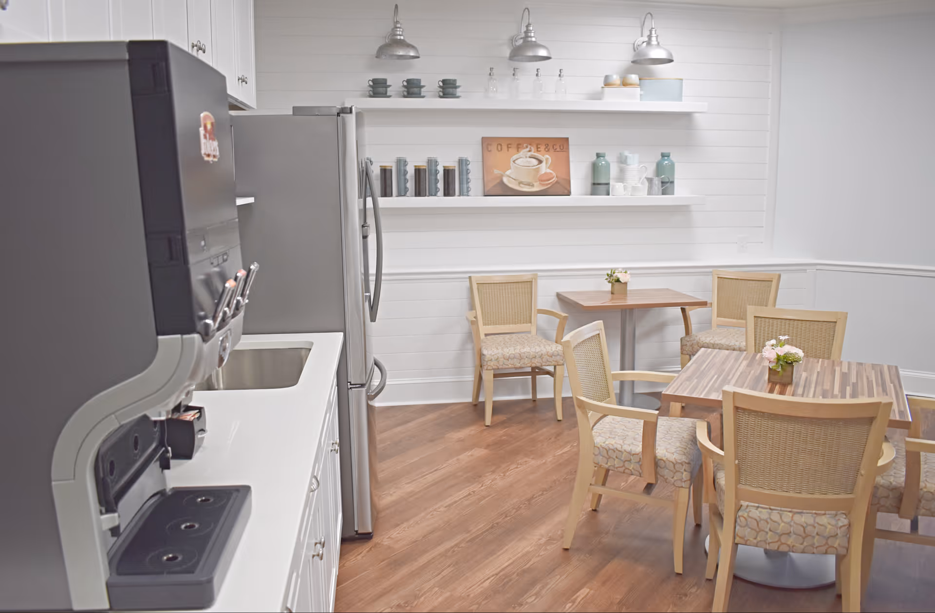 A small dining area with two wooden tables and six cushioned chairs. The room has white walls with horizontal paneling and two white shelves holding cups, jars, and decorative items. On the left side, there is a coffee machine, a stainless steel refrigerator, and a sink with white cabinetry below. The floor is wood with a warm tone.