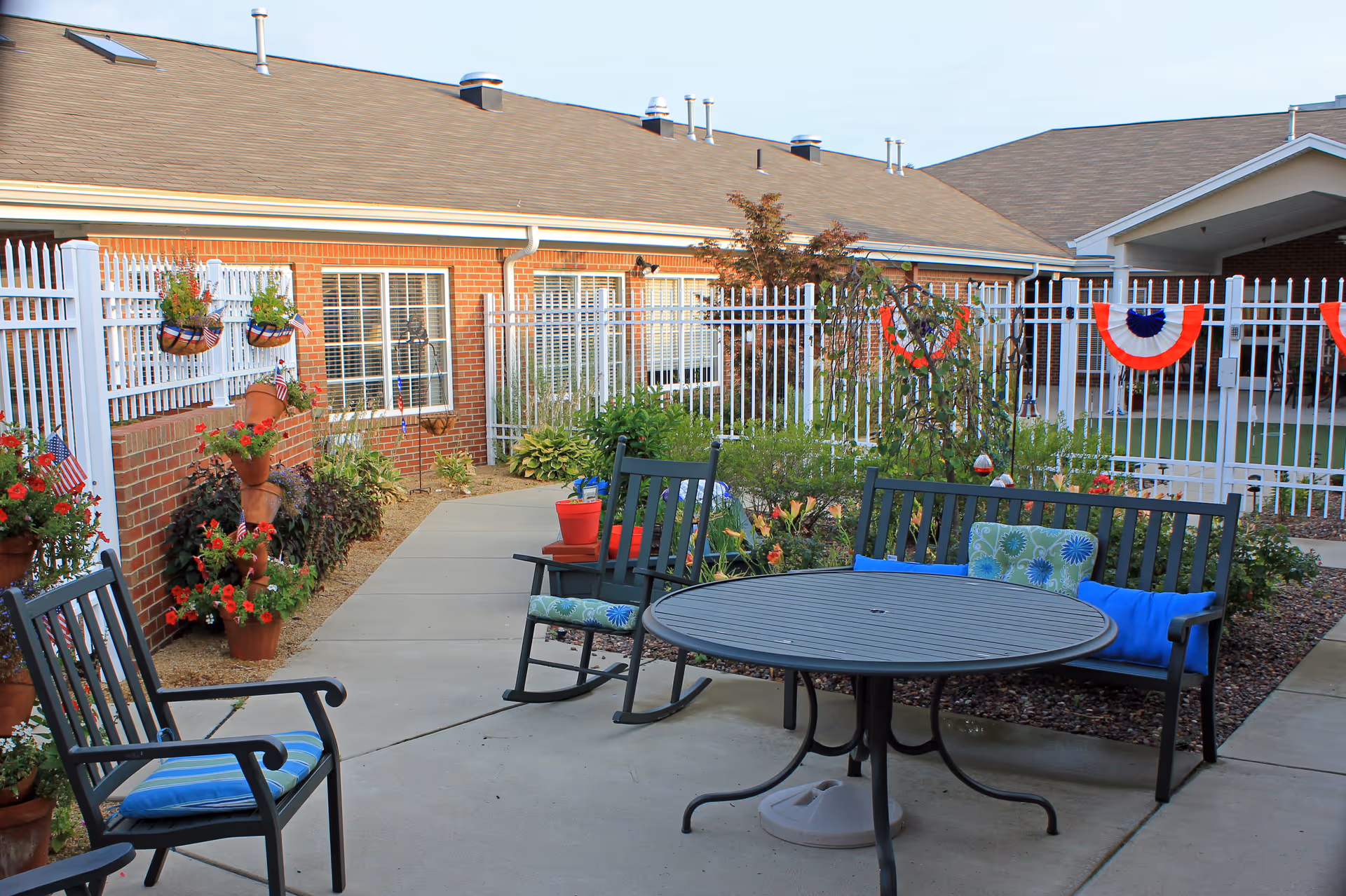Outdoor patio area at Springhurst Health Campus with black metal chairs and a round table. The chairs have blue and green cushions. The patio is surrounded by a white metal fence decorated with red, white, and blue bunting. There are various potted plants and flowers along the brick wall and garden beds.