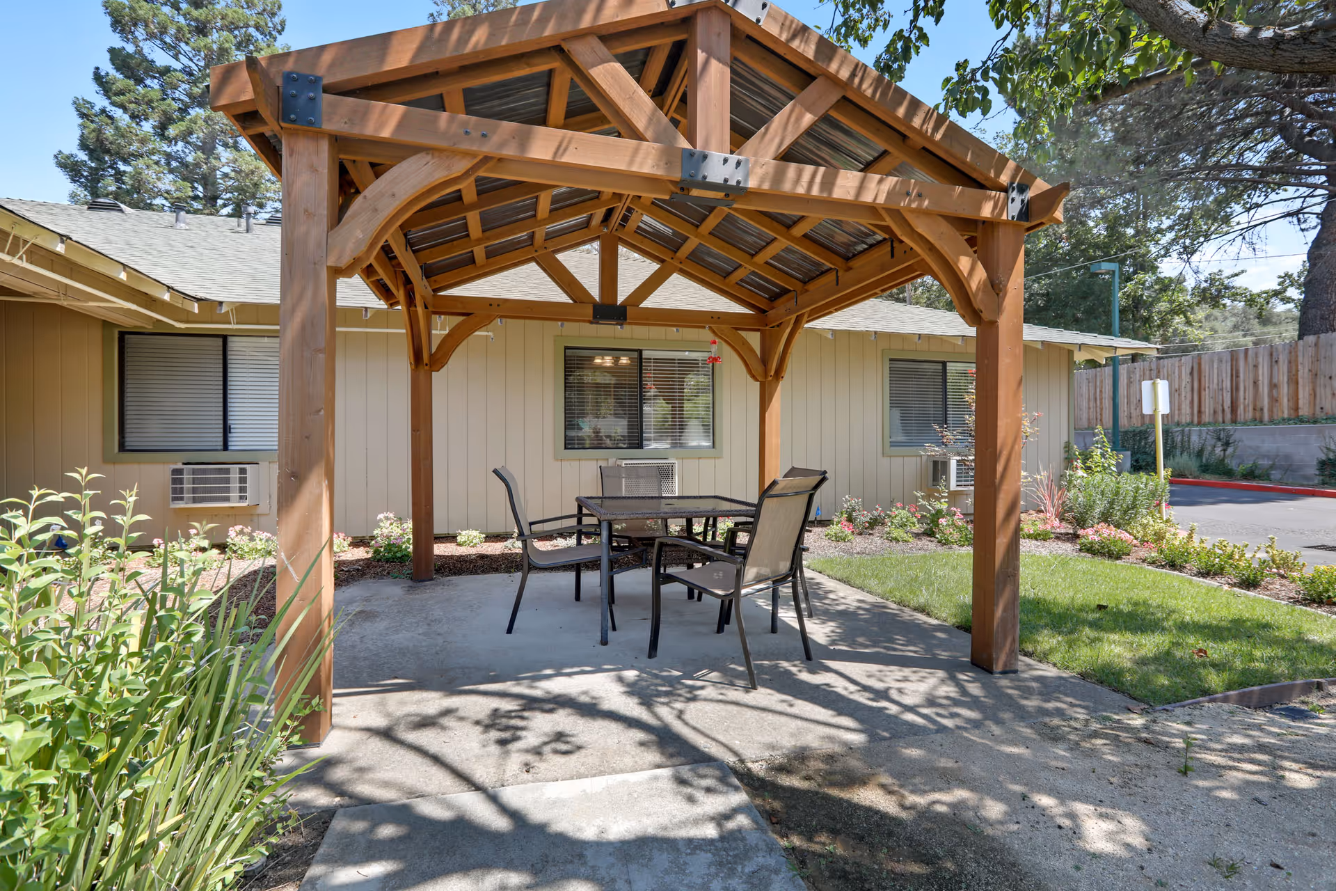 Outdoor wooden gazebo with a metal roof covering a glass-top table and four chairs, situated on a concrete patio in front of a beige building with windows and air conditioning units. Surrounding the patio are green grass, plants, and flowers under a clear blue sky.