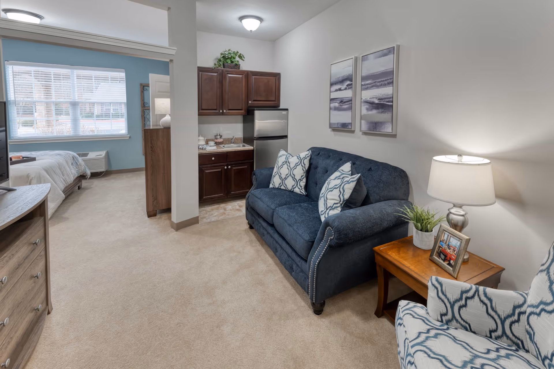 A cozy senior living apartment interior featuring a blue upholstered loveseat with patterned pillows, a wooden side table with a lamp, plant, and framed photo, a patterned armchair, a small kitchenette with dark wood cabinets and a stainless steel mini fridge, and a bedroom area with a bed and large window with blinds.