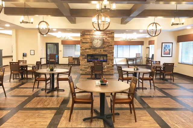 A spacious dining room with multiple round tables and wooden chairs arranged neatly. The room features a stone fireplace with a clock above it, large windows with blinds, and ceiling beams with modern light fixtures. The floor has a wood pattern with dark borders.