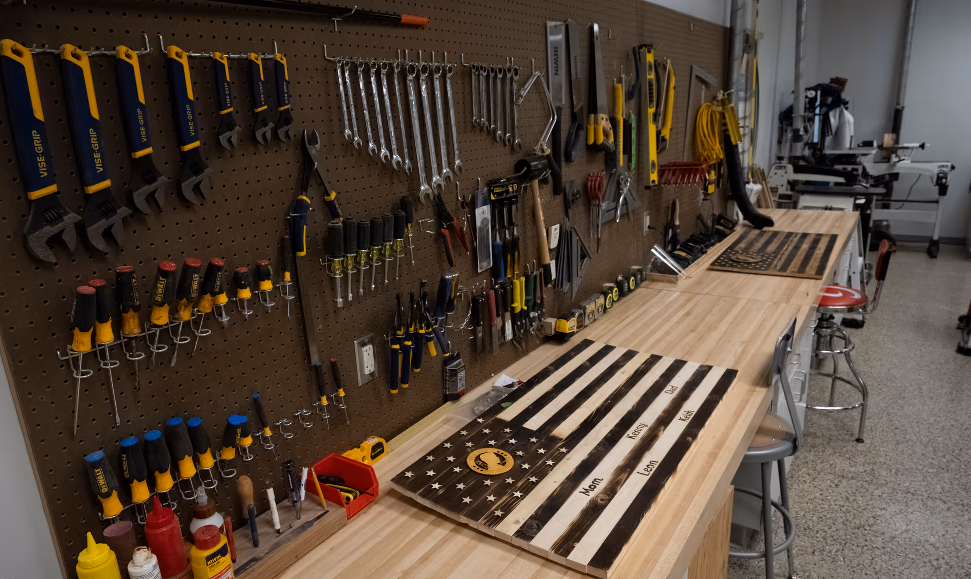 A workshop area with a wooden workbench and pegboard wall holding various tools including wrenches, screwdrivers, hammers, and measuring tapes. On the workbench are two wooden American flag plaques, one with names engraved on it. The room has stools and additional equipment in the background.