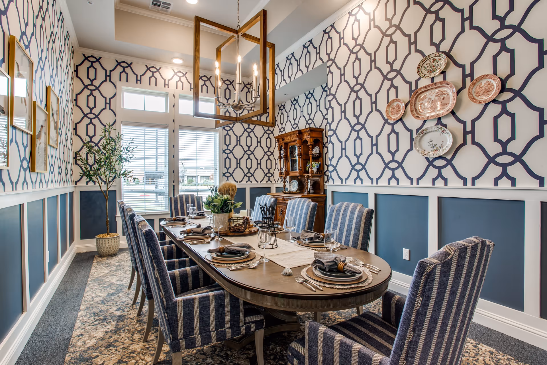 A well-decorated dining room with a long oval wooden table set for eight people. The chairs have blue and white vertical stripes. The walls feature a blue and white geometric patterned wallpaper with decorative plates mounted on one wall and framed pictures on another. A wooden cabinet with glass doors stands against the far wall. A modern chandelier with exposed bulbs hangs above the table, and a potted plant is placed near the window with white blinds.