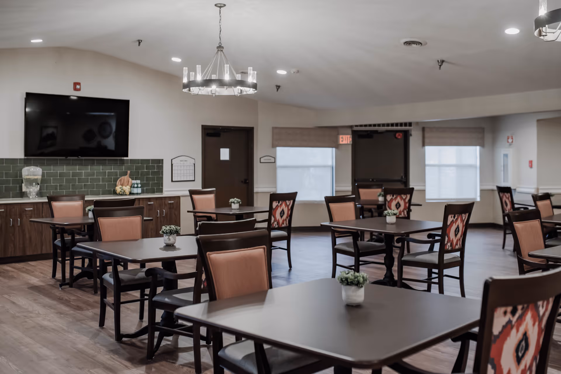 A dining room in a senior living facility with multiple tables and chairs arranged neatly. Each table has a small potted plant as decoration. There is a large flat-screen TV mounted on the wall above a cabinet with a water dispenser and decorative items. The room has wood flooring, a chandelier, and windows with blinds.