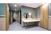 Interior hallway of a senior living facility with a wall-mounted sink and two round mirrors. The hallway has light blue walls, wooden doors on both sides, and recessed ceiling lights.