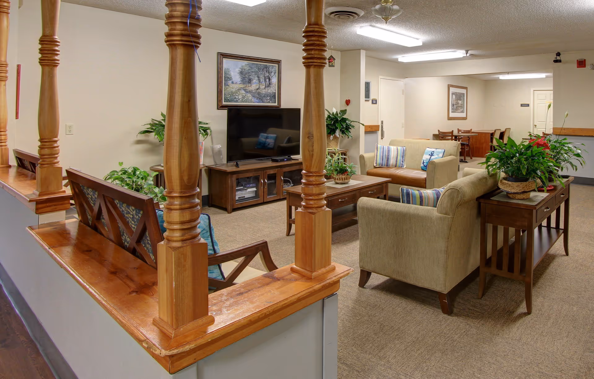 A cozy senior living common area with beige and brown upholstered sofas, a wooden coffee table with a plant, a TV on a wooden stand, and several green plants placed around the room. The space is softly lit with fluorescent ceiling lights and decorated with framed artwork on the walls. Wooden railings partially separate the seating area from the hallway.