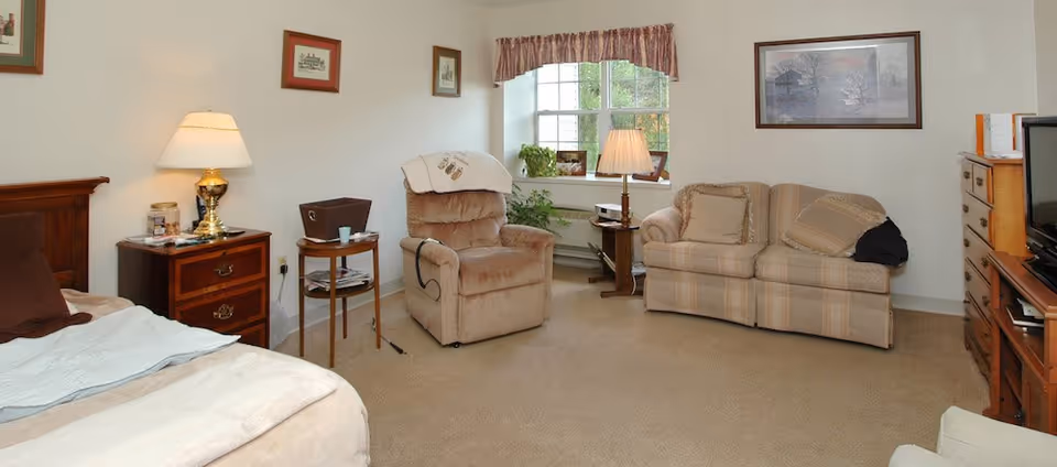 A cozy living area in a senior living facility featuring a beige recliner chair, a two-seater striped sofa with cushions, a wooden TV stand with a television, a wooden nightstand with a lamp, and a bed partially visible on the left. The room has beige carpet, framed pictures on the walls, and a window with a valance letting in natural light.