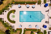 Aerial view of a rectangular outdoor swimming pool surrounded by lounge chairs, umbrellas, pathways, and landscaped greenery with a circular fountain feature nearby.