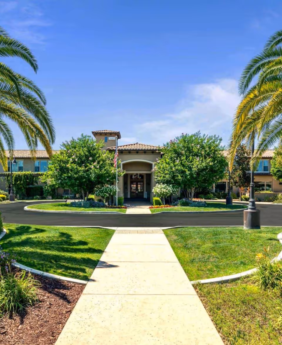 Front exterior view of Oakmont of Fresno senior living facility with a clear blue sky, palm trees, green lawns, and a paved walkway leading to the entrance.