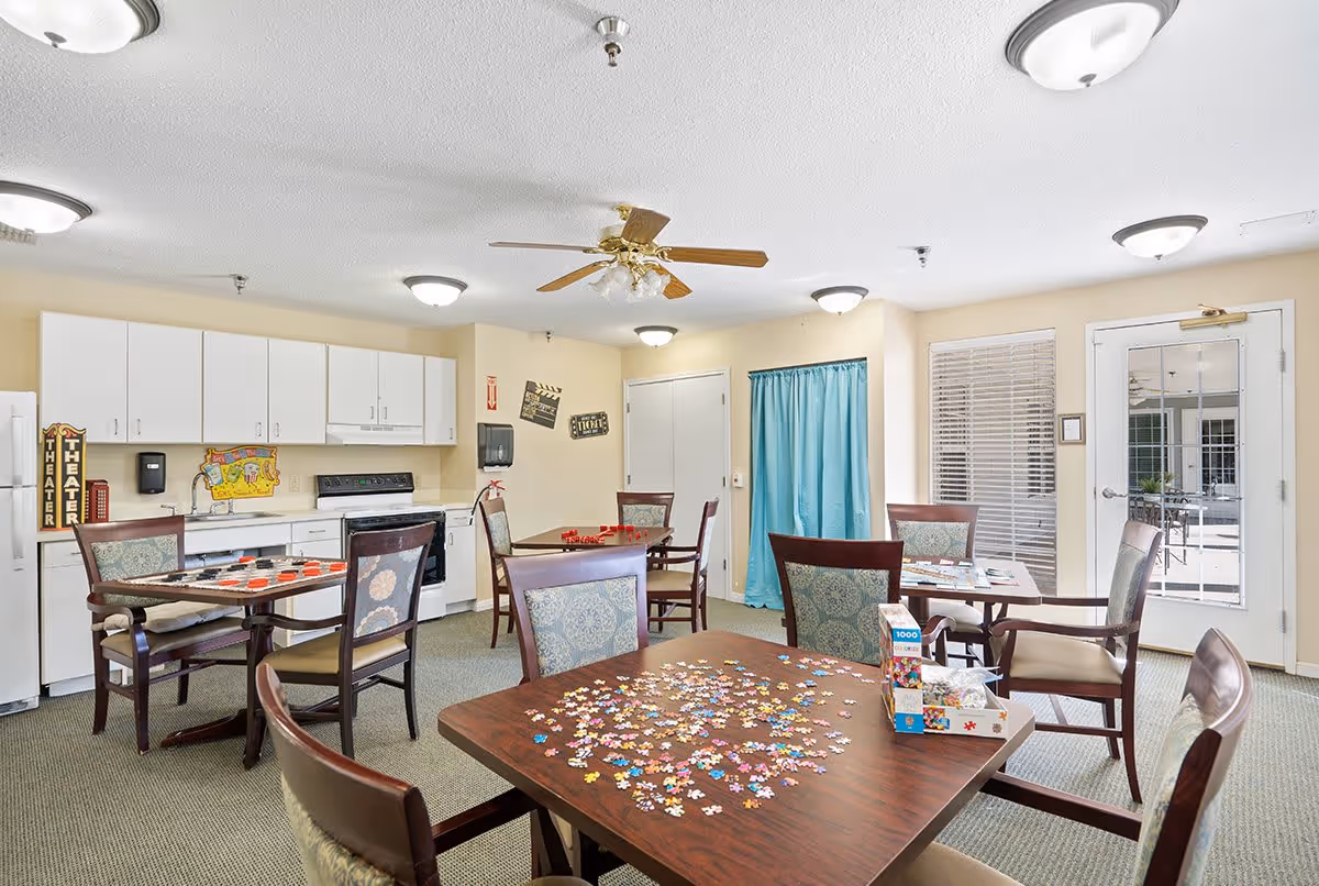 A bright activity room in a senior living facility with several tables and chairs arranged for games and puzzles. One table has a partially completed jigsaw puzzle with scattered pieces and a puzzle box. Another table has a checkers game set up. The room includes a kitchenette with white cabinets, a stove, and a refrigerator. The walls are decorated with theater-themed signs and a colorful map. There is a ceiling fan and multiple ceiling lights, a door with a window, and a window with blinds and a blue curtain.