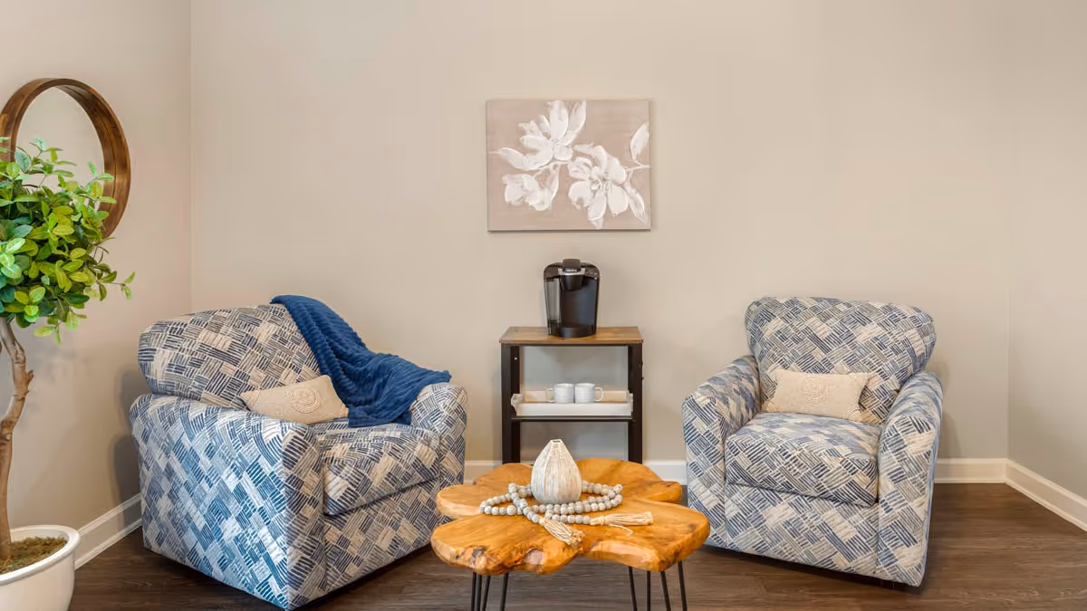 A cozy seating area with two patterned armchairs facing each other, a wooden coffee table with decorative items in between, a small table with a coffee maker and cups against the wall, a potted plant on the left, and a floral artwork hanging on the beige wall.