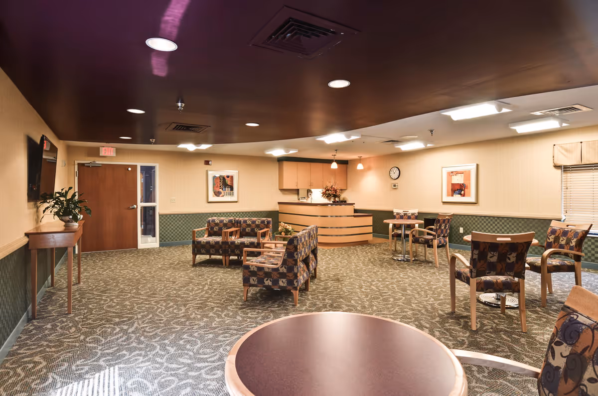 A spacious, well-lit communal lounge with patterned armchairs, small tables, and a reception desk at the far wall.