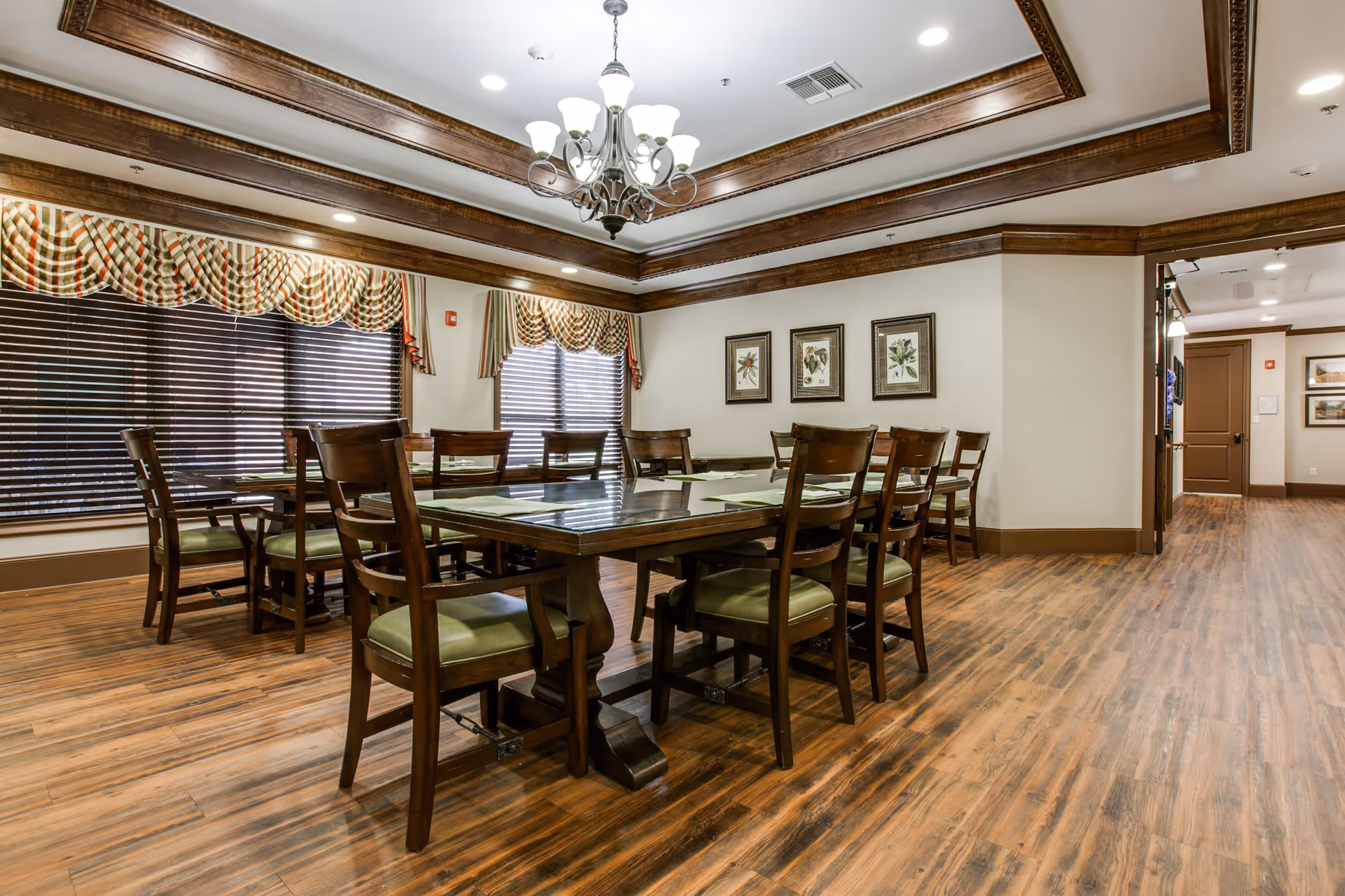 A dining room with a large wooden table surrounded by wooden chairs with green cushions. The room features wood flooring, decorative crown molding, a chandelier, and windows with patterned valances and blinds. Three framed botanical prints hang on the wall.
