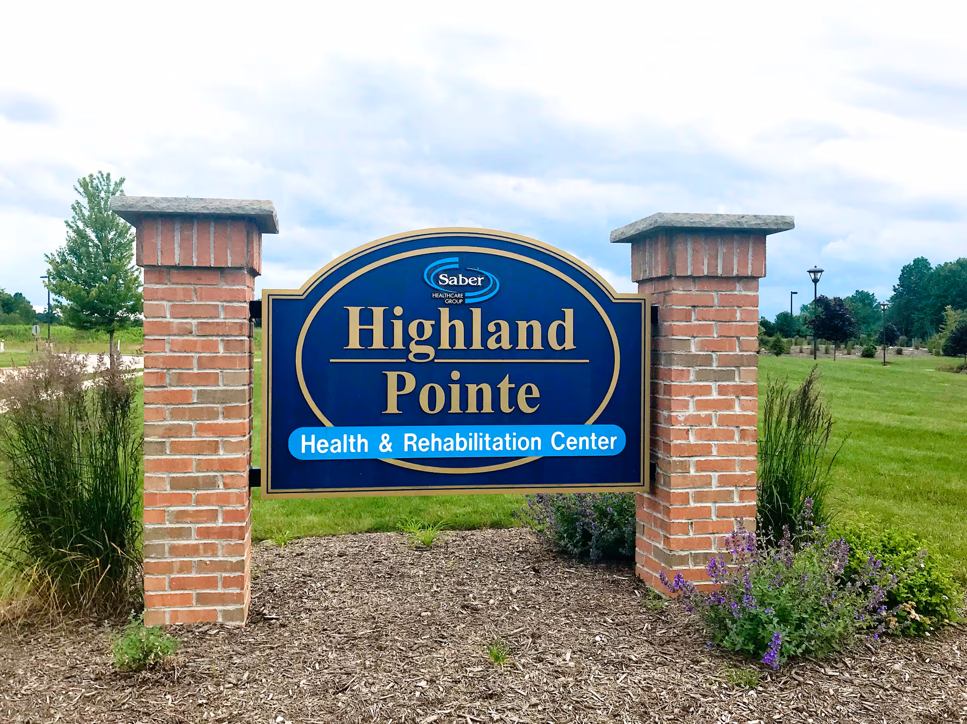 A large blue and gold sign for Highland Pointe Health & Rehabilitation Center mounted between two brick pillars, with grass, plants, and trees in the background under a cloudy sky.