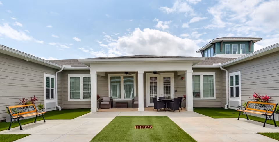 Outdoor courtyard area of a senior living facility with two benches on either side, a covered patio with seating and a table, surrounded by beige buildings with white trim and windows under a partly cloudy sky.