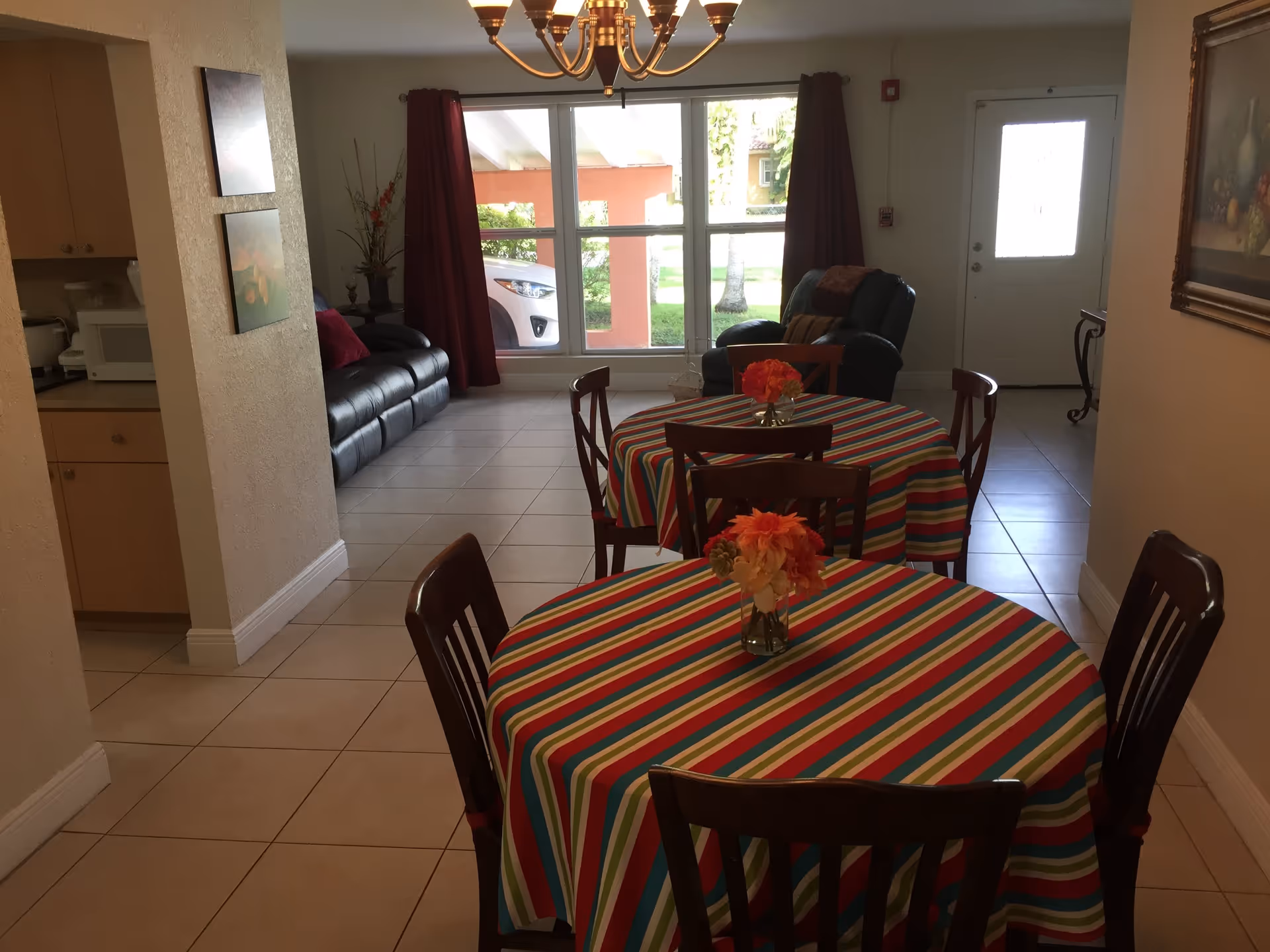 Interior view of a senior living facility dining and living area with two round tables covered in colorful striped tablecloths, each with a small flower arrangement. In the background, there is a black leather sofa, a black armchair, large windows with red curtains, and a door leading outside. A chandelier hangs from the ceiling.
