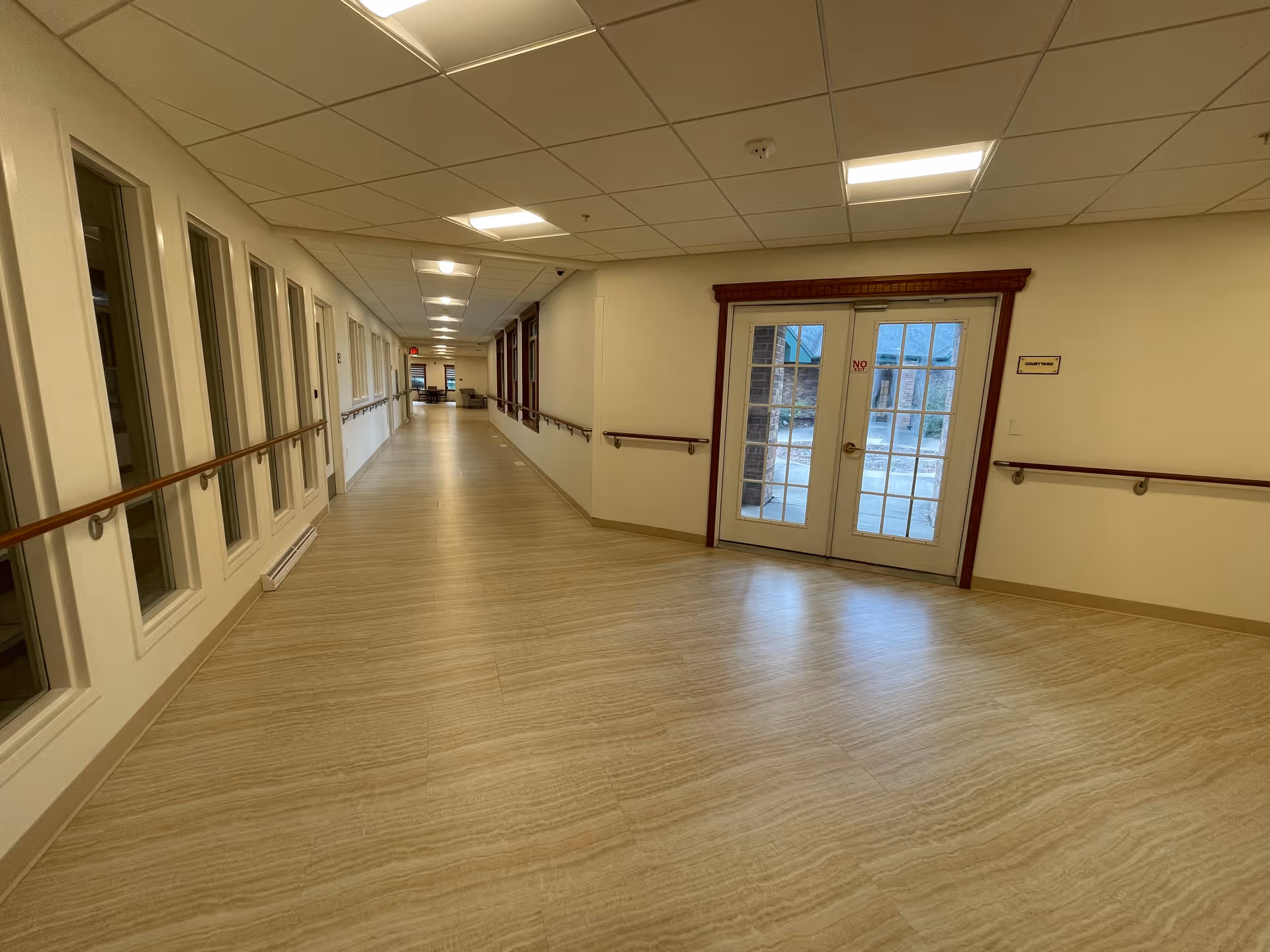 Wide empty interior hallway with handrails, windows along the left wall, and glass double doors at the far end.