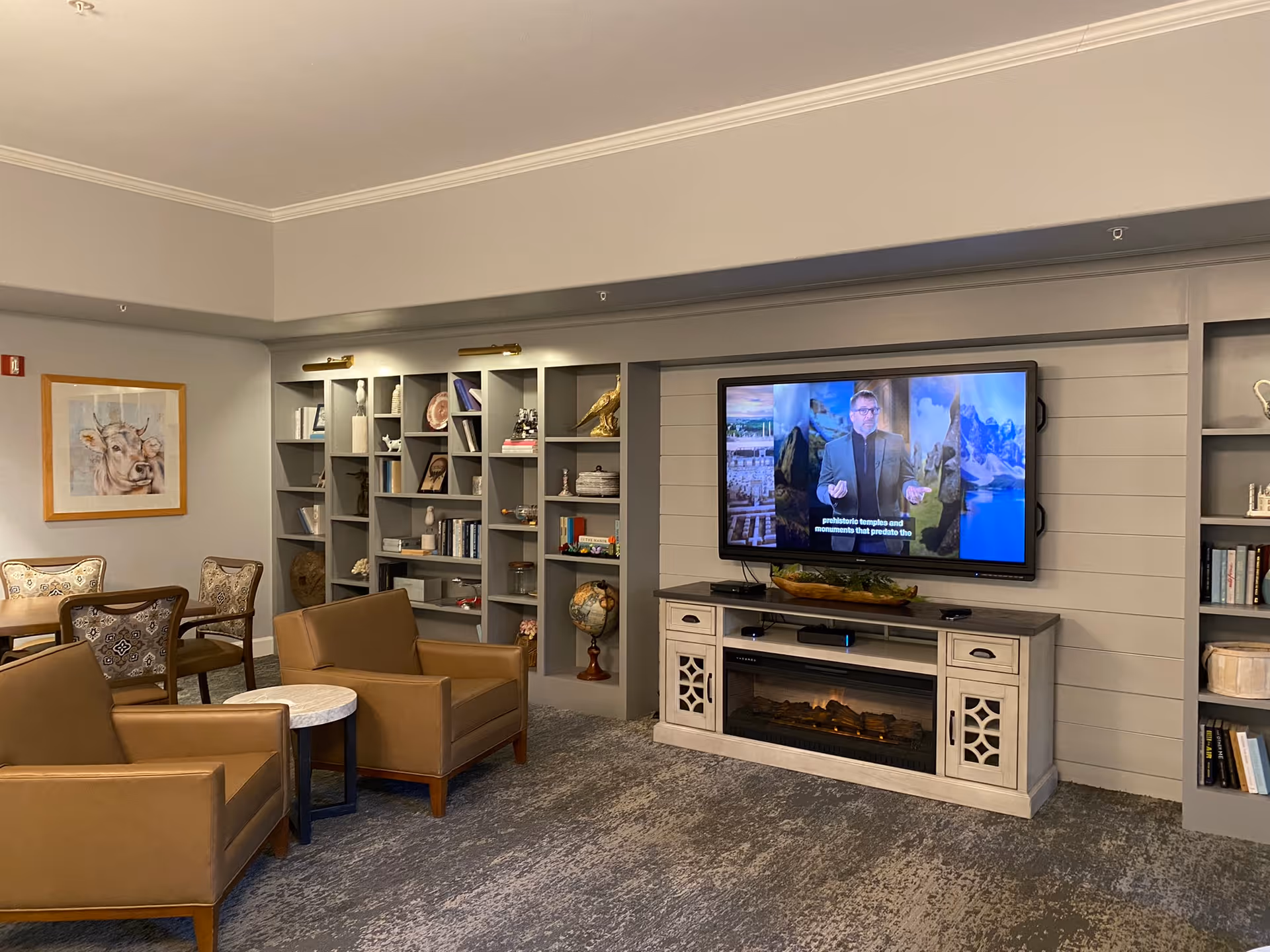 Comfortable common living area with armchairs, bookshelves, and a wall-mounted TV above a fireplace console.