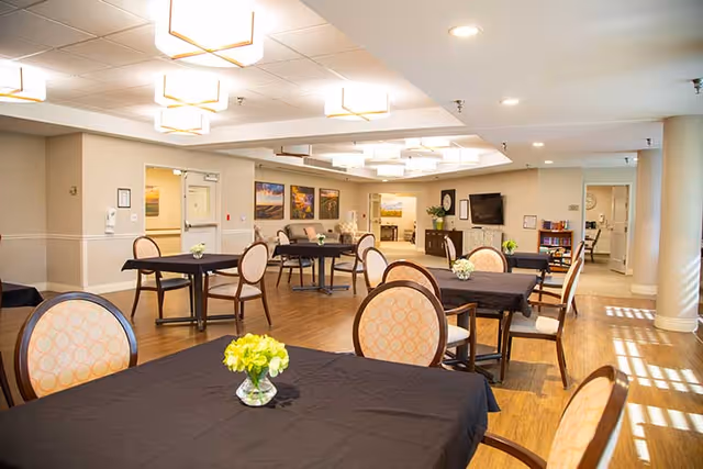 Bright communal dining/activity room with multiple tables and chairs, floral centerpieces, and a seating area with a TV.