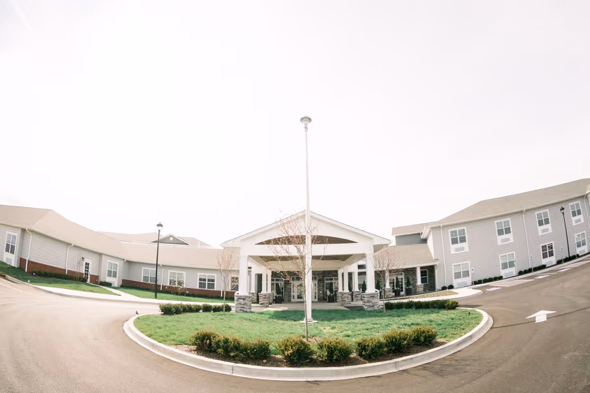 Front exterior view of Bluegrass Senior Living facility showing a large entrance with a covered drop-off area, surrounded by a circular driveway and landscaped greenery.