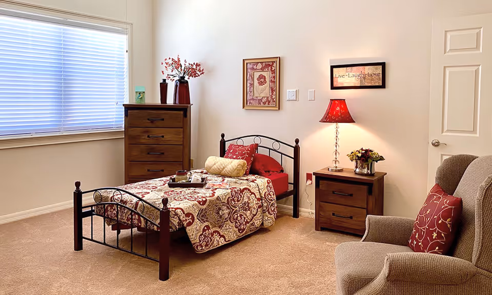 A cozy bedroom with a single bed featuring a patterned red and beige bedspread and matching pillows. Next to the bed is a wooden nightstand with a red lamp and a small flower arrangement. A wooden dresser with decorative vases and flowers is placed near a window with closed blinds. A comfortable armchair with a red patterned pillow is positioned in the corner. The walls are decorated with framed artwork and a 'Live Laugh Love' sign.
