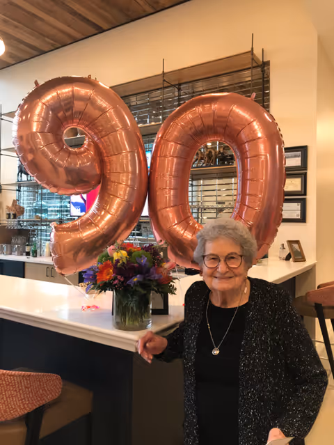 An elderly woman with gray hair and glasses smiling while standing next to a counter with a bouquet of colorful flowers and large rose gold balloons shaped as the number 90 in the background. The setting appears to be a modern indoor common area with a wooden ceiling and shelves with glassware.