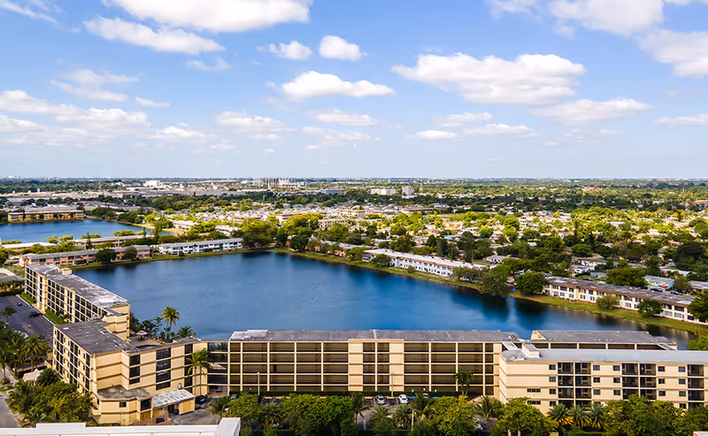 Aerial view of a senior living facility named Grand Court Lakes surrounding a large lake, with multiple beige multi-story buildings, palm trees, and a sprawling suburban area in the background under a partly cloudy blue sky.