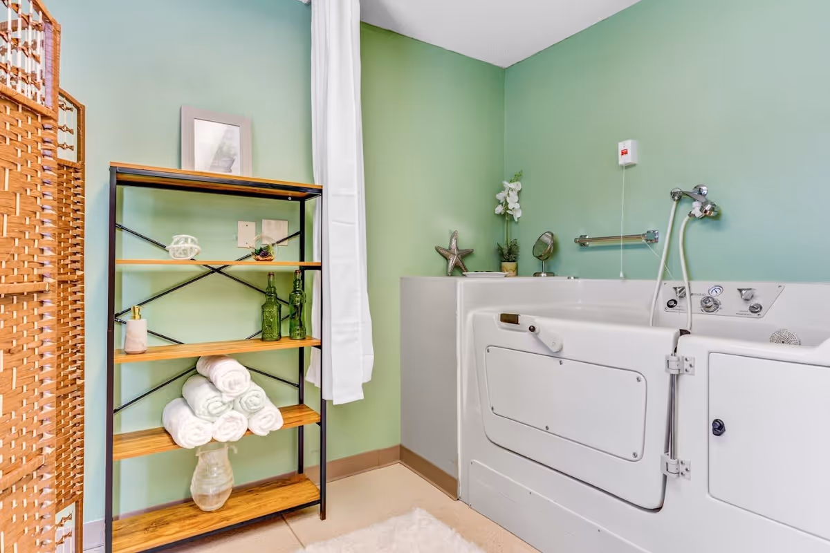 A bathroom with a walk-in bathtub featuring a door and handrails, a showerhead, and controls on the side. Next to the bathtub is a wooden shelving unit holding rolled white towels, green glass bottles, a soap dispenser, and decorative items. The walls are painted light green, and there is a white curtain hanging beside the shelving unit.