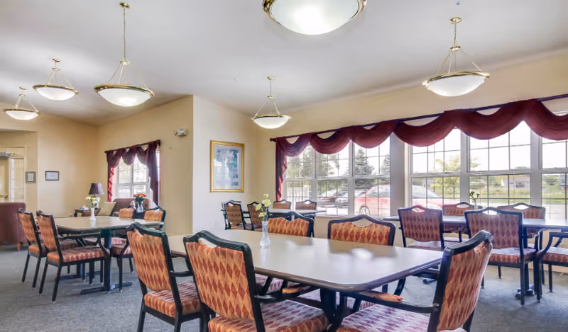 Bright communal dining room with multiple tables and patterned chairs beneath pendant lights and large windows with red valances.