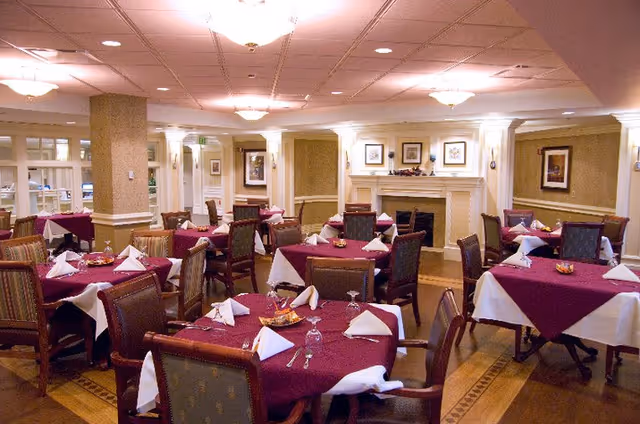 A dining room with multiple tables covered in burgundy and white tablecloths, each set with folded white napkins, glasses, and silverware. The room features wooden chairs with cushioned seats, a fireplace with framed artwork above it, and soft ceiling lighting.
