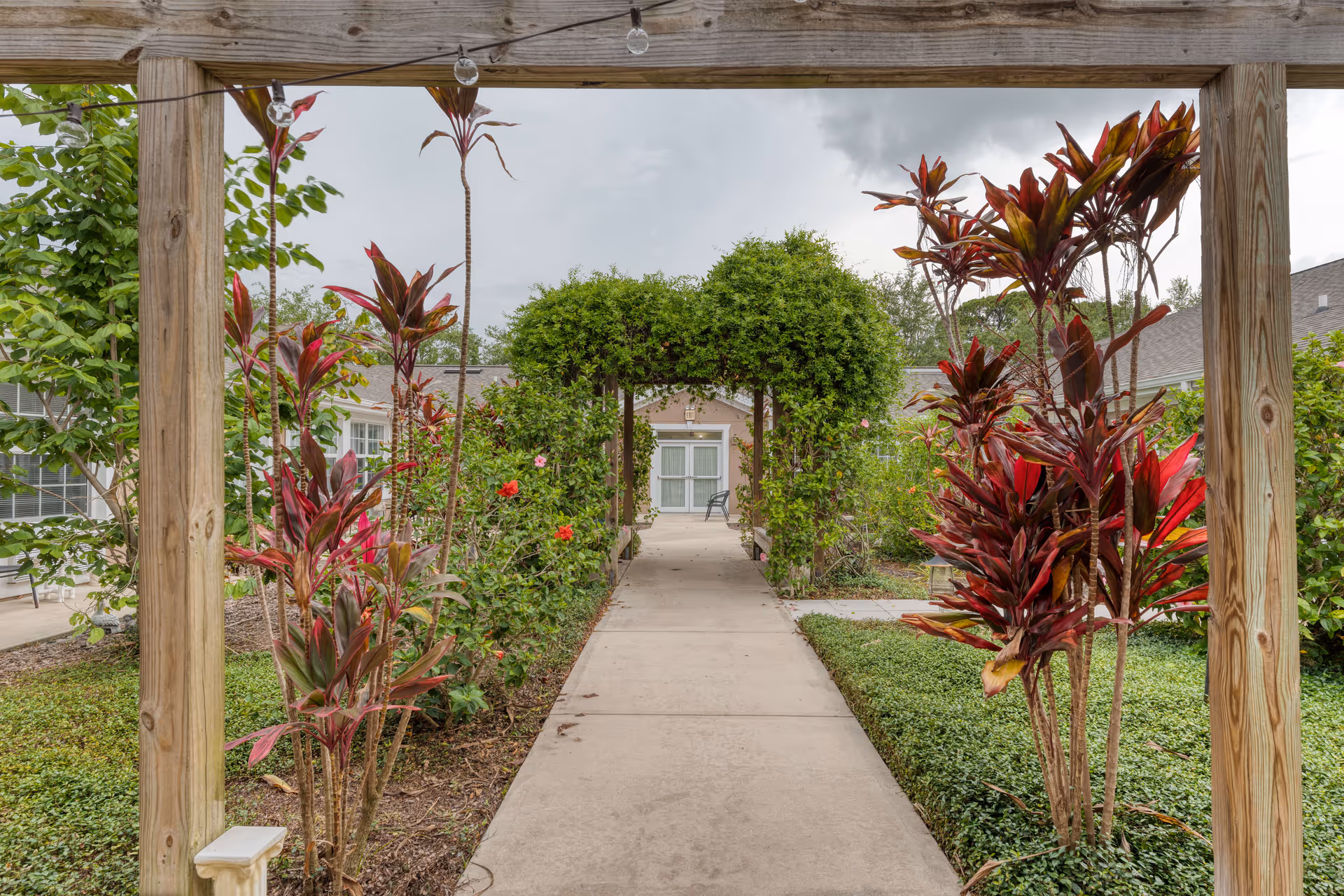 A garden pathway at Addington Place of Titusville with a wooden pergola overhead and lush greenery on both sides. The path leads to a green archway covered with climbing plants, with a building and a chair visible in the background under a cloudy sky.