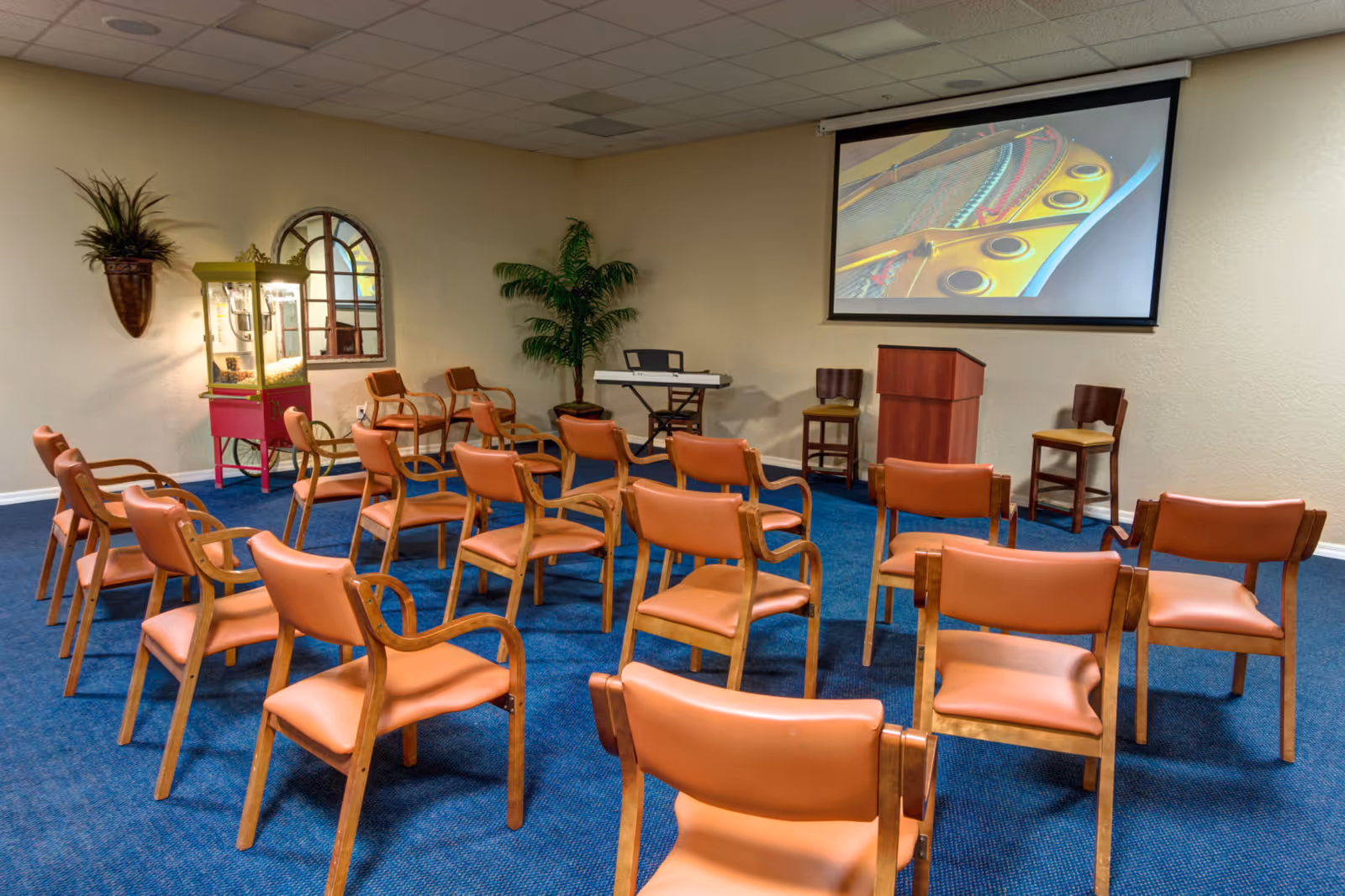 A small auditorium or meeting room with rows of orange cushioned chairs arranged facing a podium and a projection screen displaying a close-up image of a piano's interior. The room has blue carpet, beige walls, a keyboard on a stand, a popcorn machine, a wall-mounted plant, and a large potted plant in the corner.