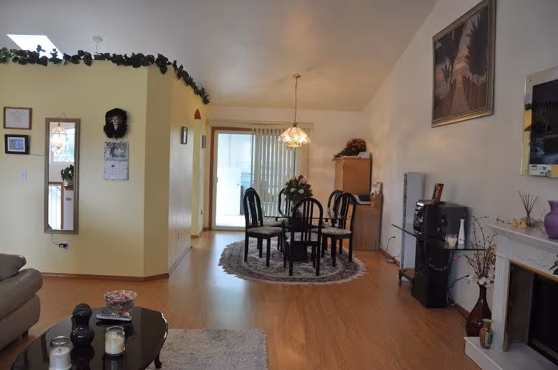 A cozy living and dining area with wooden flooring. The dining area features a round glass table with four black chairs on a circular rug, illuminated by a hanging chandelier. The living area includes a beige sofa and a black coffee table with candles and decorative items. The walls are decorated with framed pictures, a mirror, and a painting. There is a sliding glass door with vertical blinds leading to an outdoor space.
