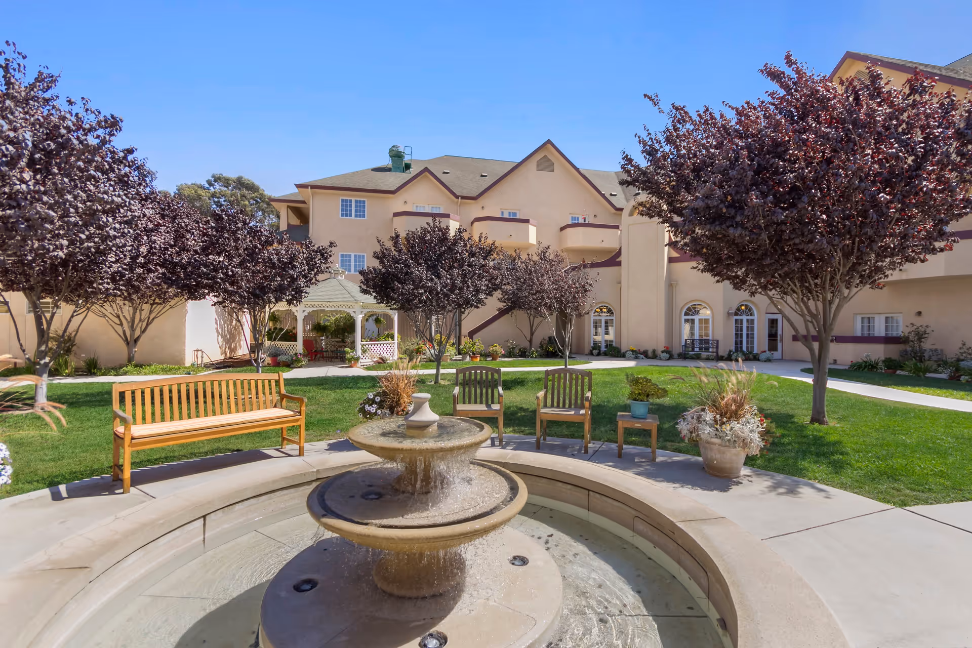 Outdoor courtyard area of a senior living facility with a central stone fountain surrounded by a curved stone bench. There are wooden benches and chairs on a grassy lawn with several purple-leaved trees. In the background, there is a beige multi-story building with balconies and arched windows, and a white gazebo.