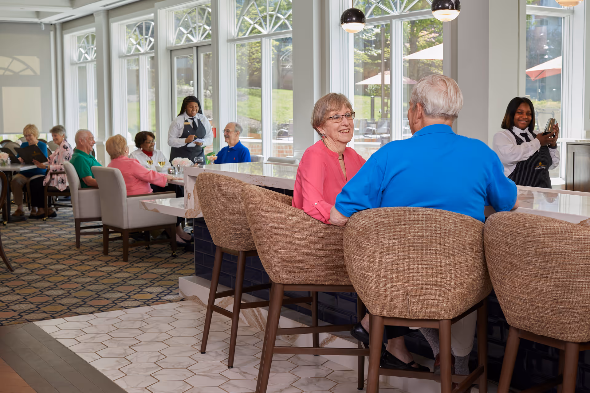 A bright dining area in a retirement community with large windows letting in natural light. Several elderly people are seated at tables and a counter, engaging in conversation and enjoying drinks. Two staff members in uniforms are attending to the residents, one taking an order and the other smiling while holding a tray.