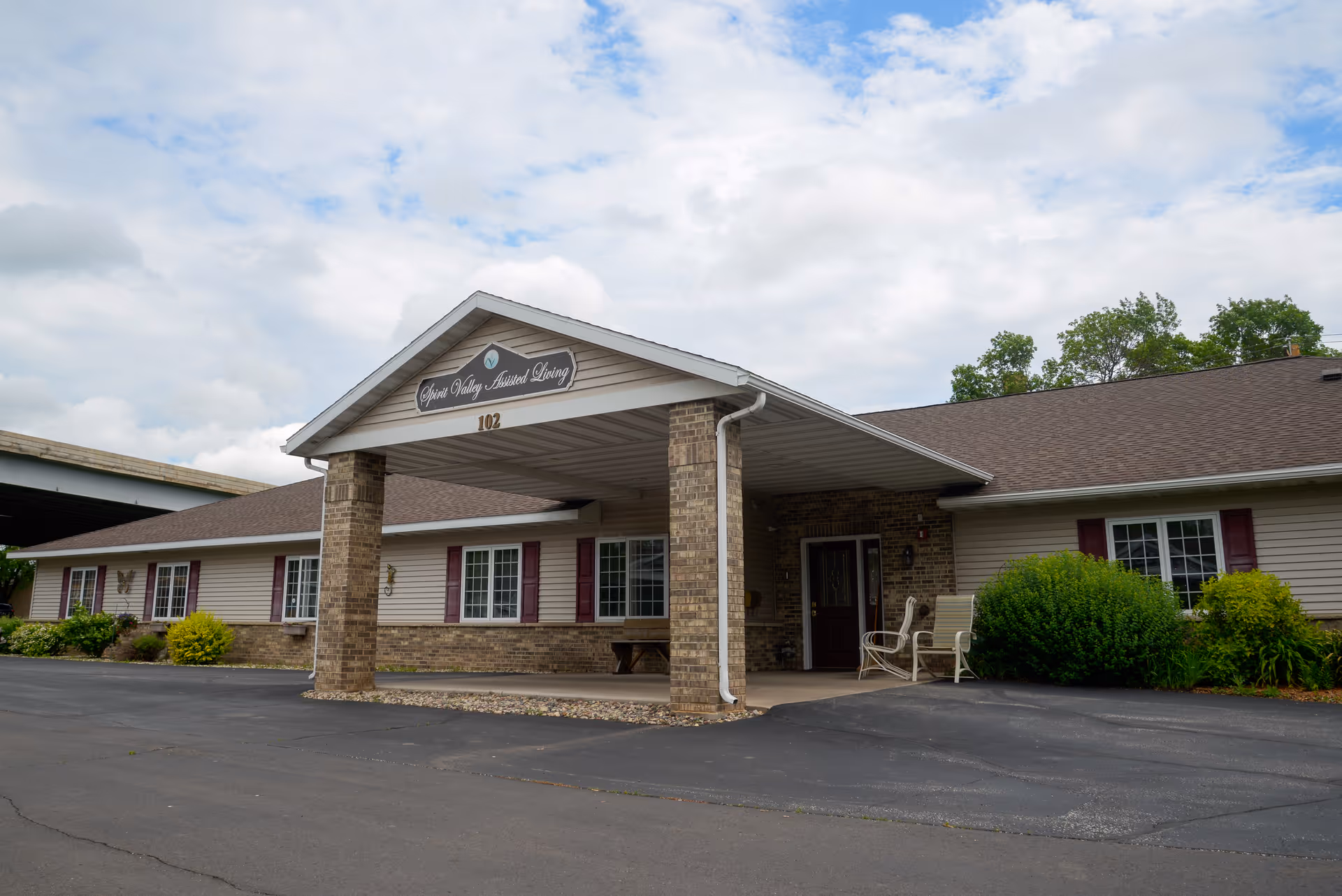 Exterior view of a single-story assisted living facility building with a covered entrance supported by brick columns. The building has beige siding, brown roof shingles, and several windows with maroon shutters. There are some bushes and greenery near the entrance and along the building. The sky is partly cloudy.