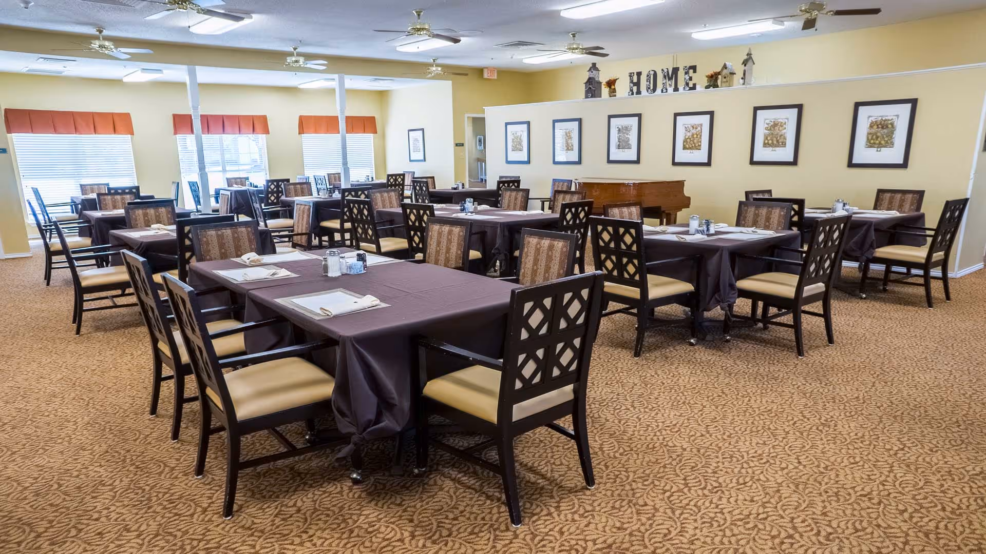 A spacious dining room with multiple tables covered in dark tablecloths, each set with napkins and condiments. The room has beige walls adorned with framed artwork and decorative letters spelling 'HOME'. Large windows with red valances let in natural light, and ceiling fans are mounted overhead. The carpet has a patterned design, and there is a piano against one wall.