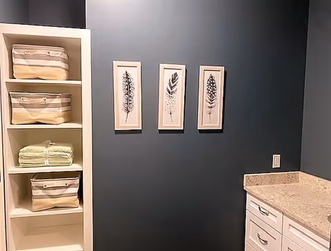 A dark teal-walled interior with white shelving holding baskets and towels, three framed feather prints on the wall, and a white countertop with drawers.