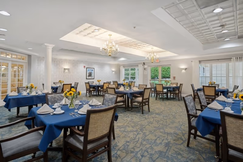 A bright and spacious dining room with multiple tables covered in blue tablecloths, each set with white napkins, glassware, and small vases of sunflowers. The room features patterned carpet, chandeliers, large windows with white curtains, and light-colored walls with subtle wallpaper. There are wooden chairs with cushioned seats around the tables.