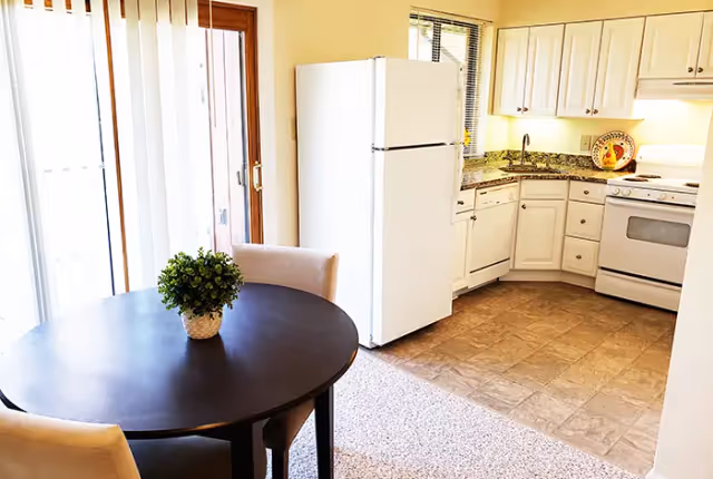 A bright kitchen and dining area featuring white cabinets, a white refrigerator, a stove, and a dishwasher. A round black dining table with two beige chairs and a small green potted plant is positioned near sliding glass doors with vertical blinds.