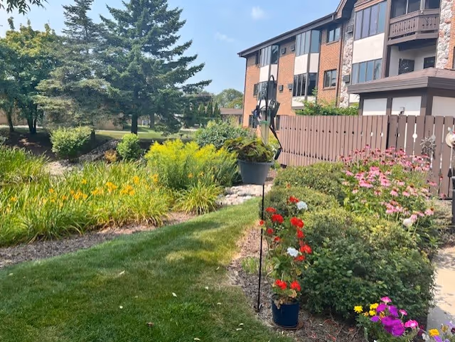 A landscaped garden area with green grass, colorful flowers including red, pink, yellow, and purple blooms, and various shrubs. In the background, there is a multi-story building with balconies and large windows. Tall trees and a clear blue sky are also visible.