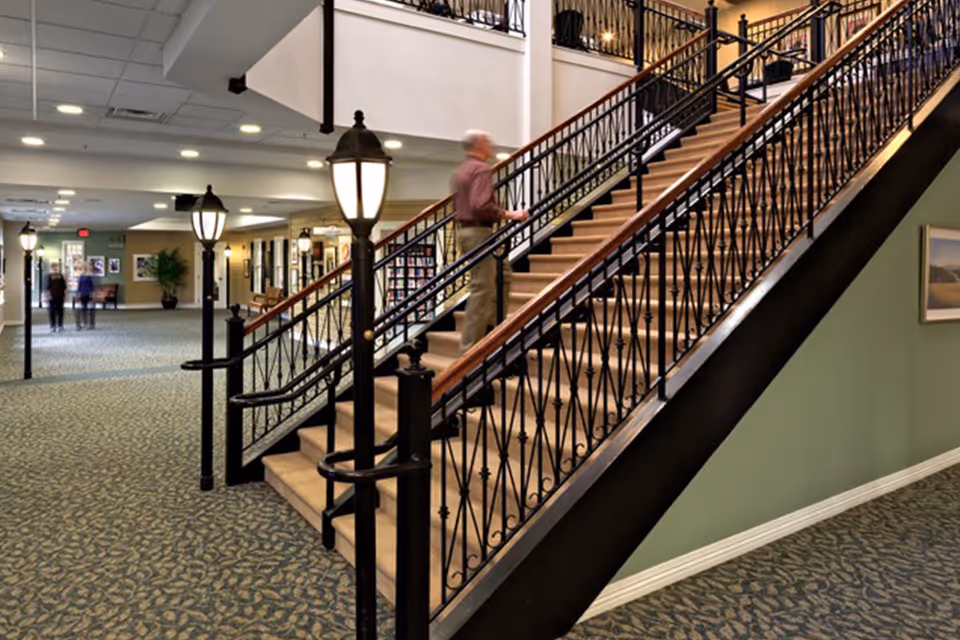 Interior view of a senior living facility hallway with carpeted floors, decorative black metal stair railings, and vintage-style lamp posts lining the walkway. A person is walking up the stairs, and a few other people are visible in the background near the hallway entrance.