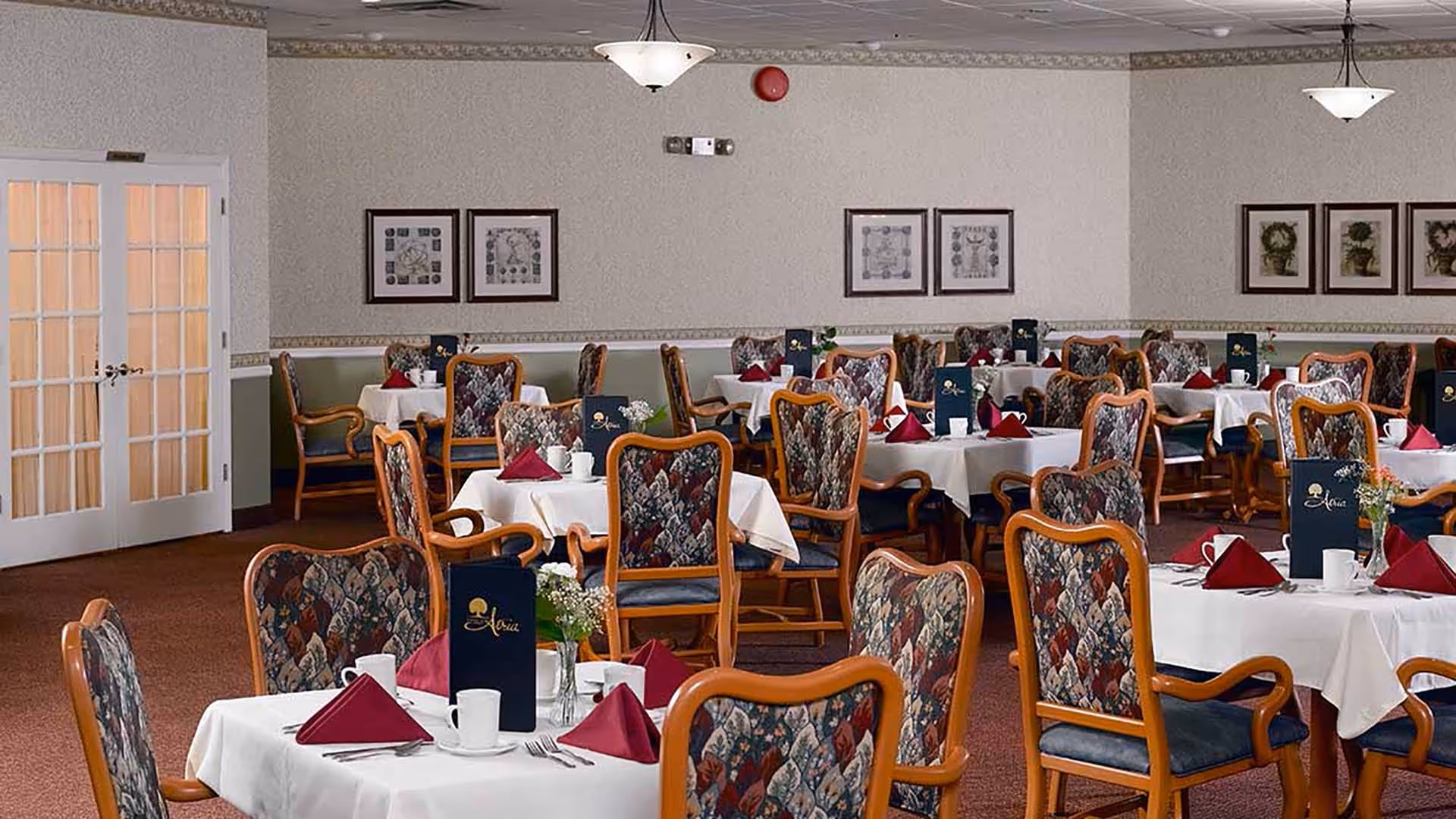 A dining room with multiple tables covered in white tablecloths, each set with folded red napkins, white cups, silverware, and small flower arrangements. The chairs have wooden frames with patterned upholstery. The room has beige walls adorned with framed artwork and two ceiling light fixtures. French doors are visible on the left side of the room.