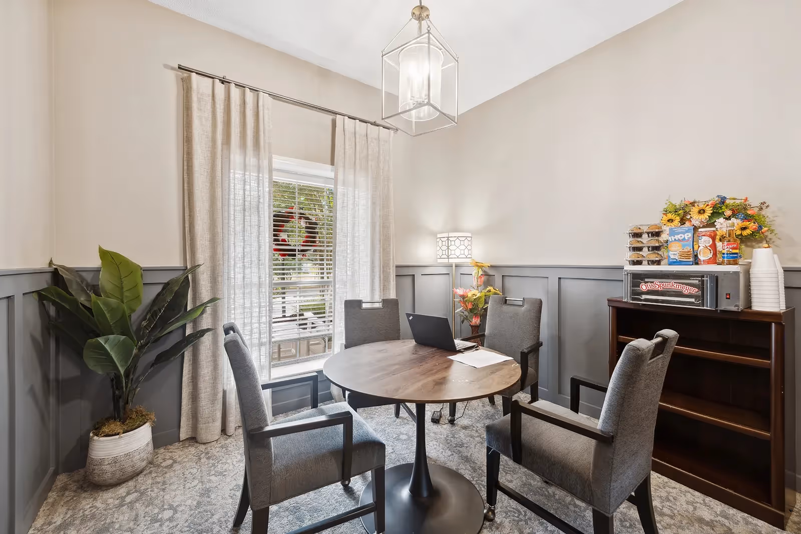 A small meeting or dining area with a round wooden table surrounded by four gray upholstered chairs. A laptop and some papers are on the table. Behind the table is a window with beige curtains and a holiday wreath visible outside. To the right, there is a dark wooden shelving unit holding a toaster oven, snacks, disposable cups, and a flower arrangement. A potted plant is placed in the corner to the left, and a floor lamp with a patterned shade stands near the shelving unit. The walls are painted beige with gray wainscoting, and a modern pendant light hangs from the ceiling.