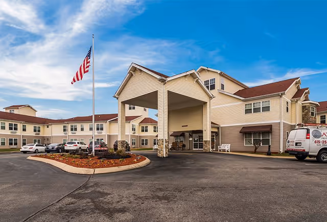Exterior view of a senior living facility named Fieldstone Place, showing a large two-story building with beige siding and red roofs. There is a covered entrance with stone pillars, a circular driveway with parked cars, a flagpole with an American flag, and a clear blue sky with some clouds.