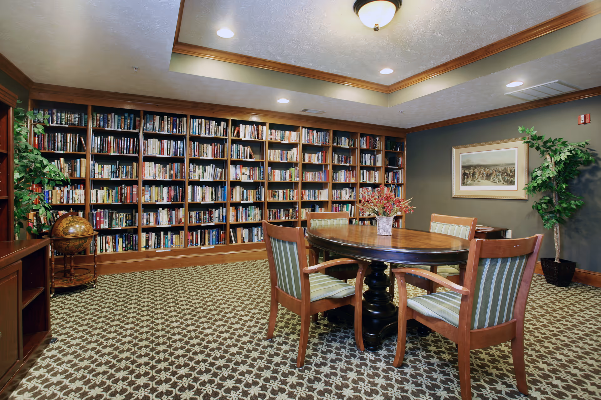 A cozy library room with a large wooden bookshelf filled with books along the back wall. In the center, there is a round wooden table surrounded by four wooden chairs with green striped cushions. The room has patterned carpet flooring, a framed picture on the wall, and two potted plants in the corners. The ceiling has recessed lighting and a central light fixture.