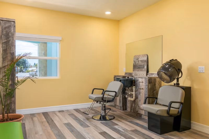 A small salon room with two styling chairs, a hooded hair dryer, a wash basin and mirror against a yellow wall.